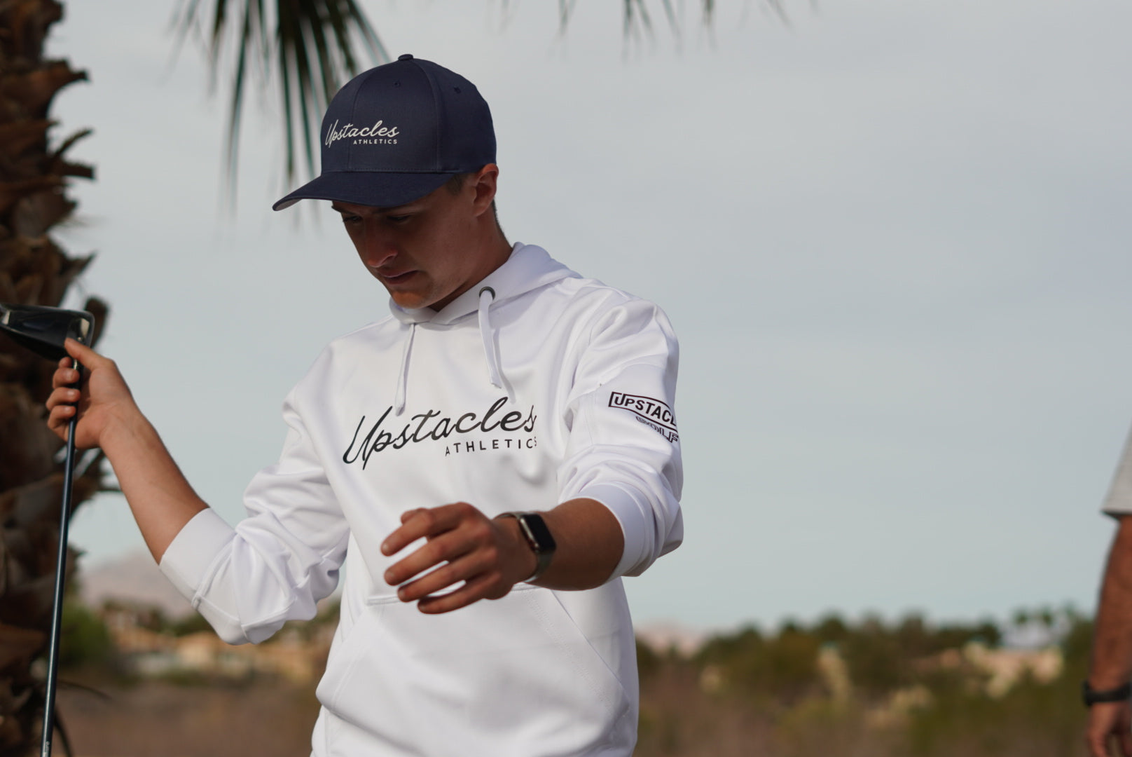 Person wearing a white hoodie and navy cap with 'Upstacles' branding, holding a golf club.